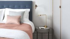 A white bedroom with a large blue bed, with white bedding, and peach blankets and cushions, white walls, and a gold lamp on a circular nightstand.
