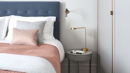 A white bedroom with a large blue bed, with white bedding, and peach blankets and cushions, white walls, and a gold lamp on a circular nightstand.