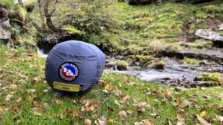 A sleeping bag in its sack, lying on grass in front of a small stream