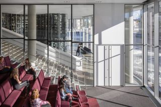 Viewing gallery, red colour seating, visitors sat in seats, glass walls looking out onto the stairwell, sun light coming into the gallery casting shadows