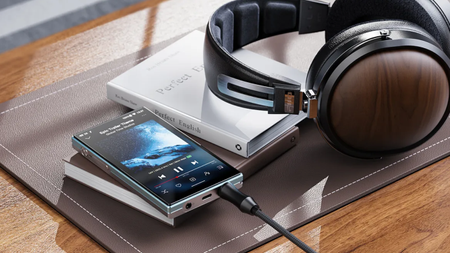 FiiO JM21 2026 on a leather mat, which in turn is on a wooden table or desk. A book and a pair of headphones sit to the right of it