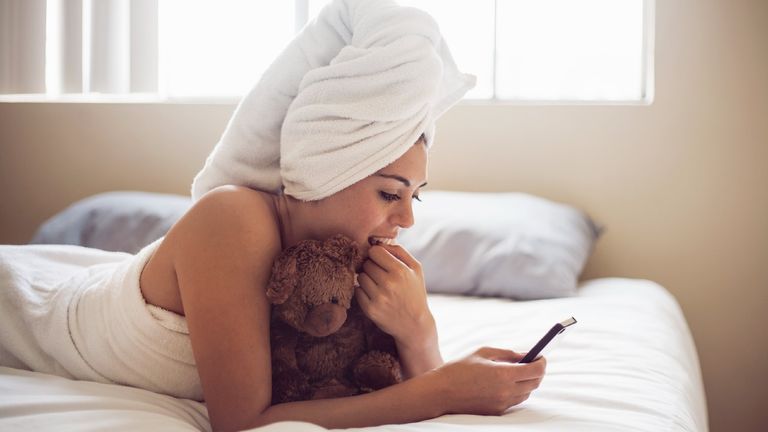 millennial woman wrapped in towel using smart phone while lying on bed holding teddy at home, millennials sleeping with teddy bears