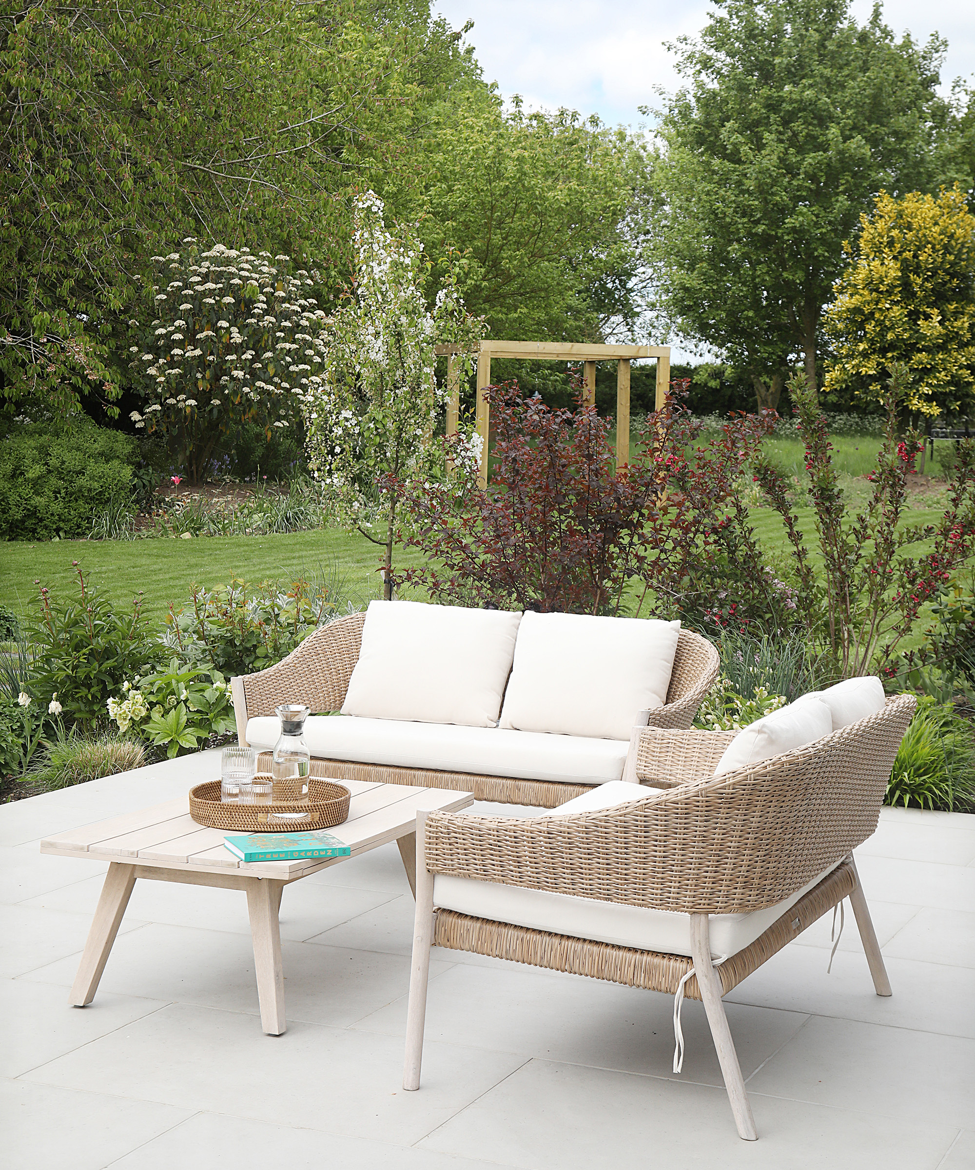 A rattan garden sofa set with coffee table and white cushions on a light patio with landscaped garden background