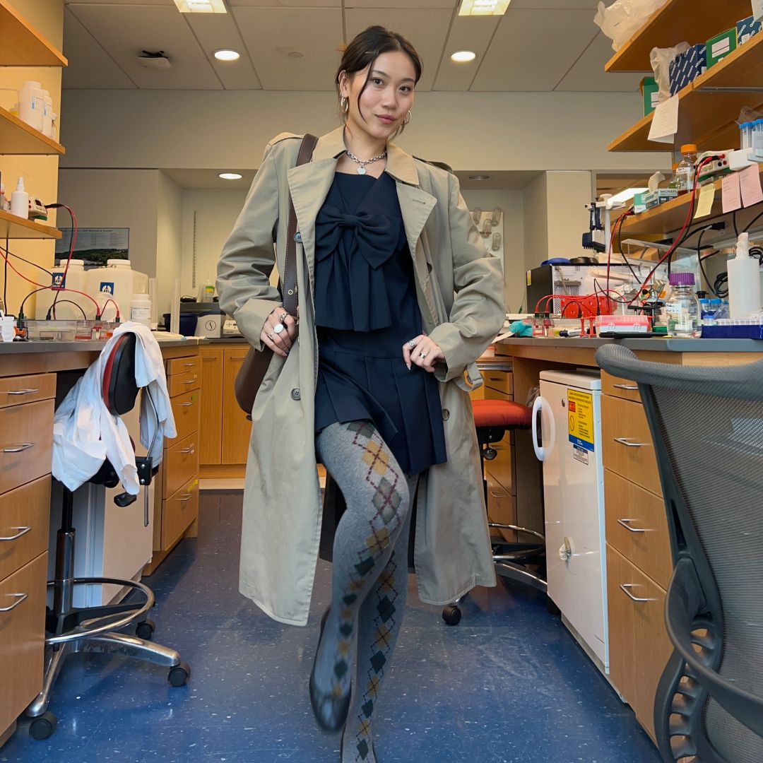 A photo of Vivian Li in a science lab wearing a black dress, argyle tights, a trench coat, and black ballet flats