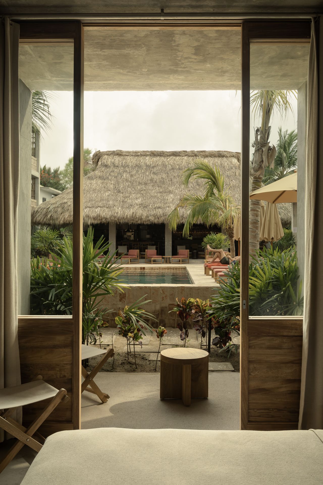 A tropical hotel features wood and rattan-clad rooms overlooking a Mexican-style pool encircled by lush trees and terracotta loungers.