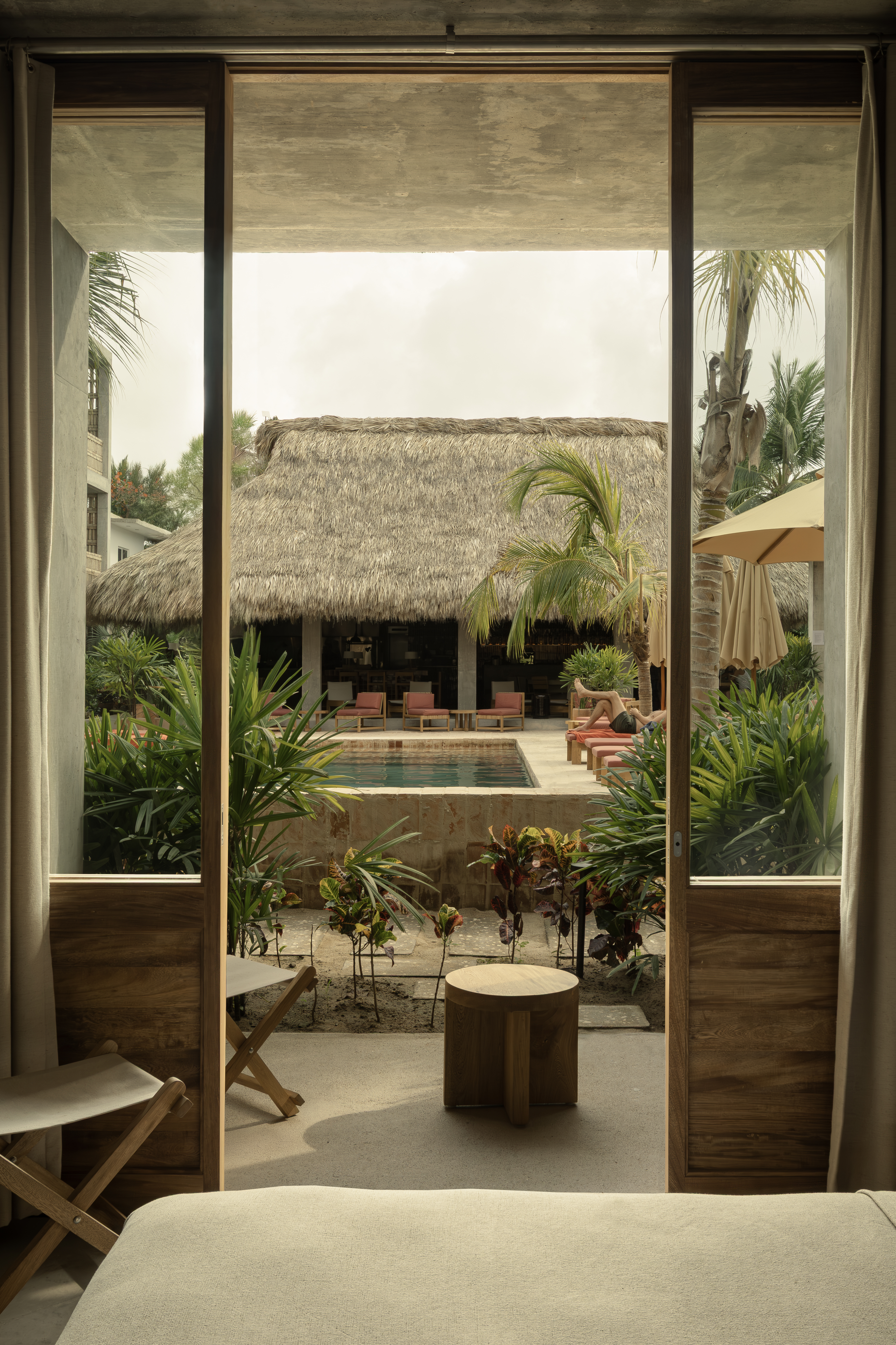 A tropical hotel features wood and rattan-clad rooms overlooking a Mexican-style pool encircled by lush trees and terracotta loungers.