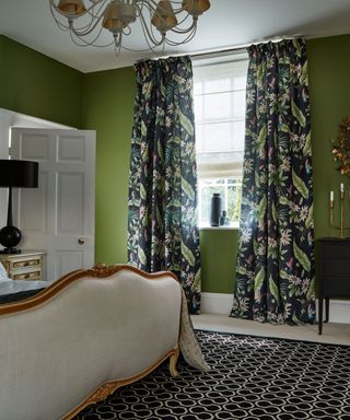 Traditional style bedroom with large bed, green painted walls, shade chandelier, black and cream rug, black chest of draws with golden rounded mirror above, tropical printed curtains
