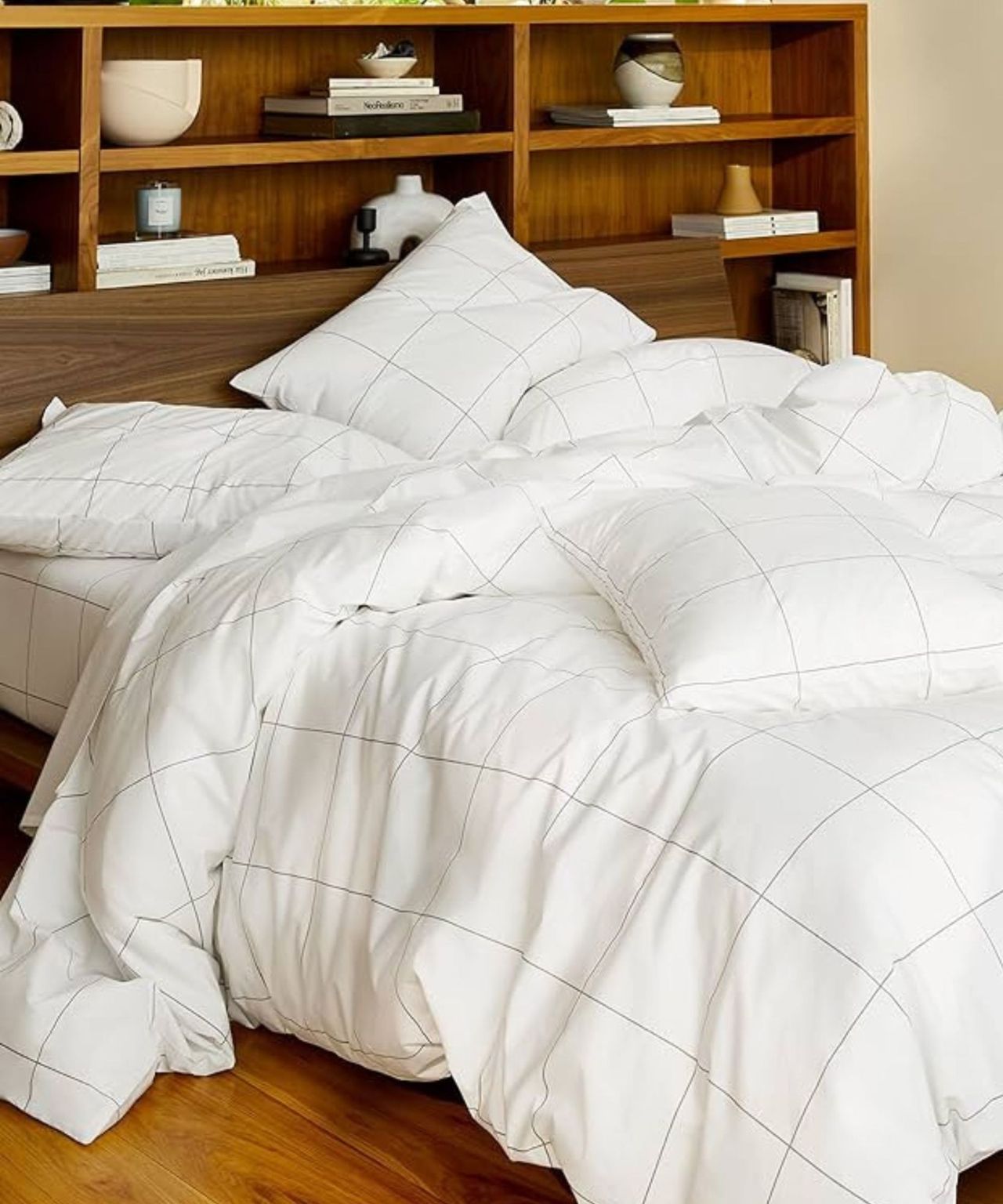 An unmade bed with a Brooklinen down comforter in a white and black grid-patterned white duvet cover, with matching pillows. The floor is a warm, polished wood, and behind are large shelves in a matching wood finish, with various vases and stacks of books.