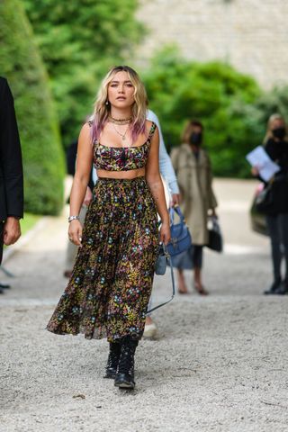 Florence Pugh wears gold pendant earrings, a gold J'Adior chain necklace, a multicolored pearls necklace, gold necklace, a black tulle with multicolored flowers embroidered zipper tank-top / crop-top, a matching black tulle with multicolored flowers embroidered flowing midi skirt, a blue gray small Lady D-Lite hand bag from Dior, a silver watch, a gold ring, black shiny leather laces knees boots from Christian Dior, outside Dior, during Paris Fashion Week - Haute Couture Fall/Winter 2021/2022, on July 05, 2021 in Paris, France