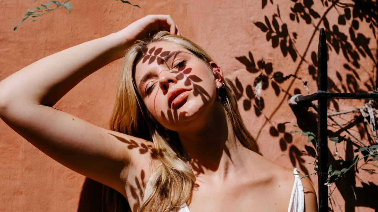 woman with blonde hair leaning against a wall with shadows of leaves on her face