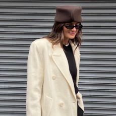 Coco Schiffer wears a brown pill-box hat, white coat, black sweater and black, oval sunglasses.