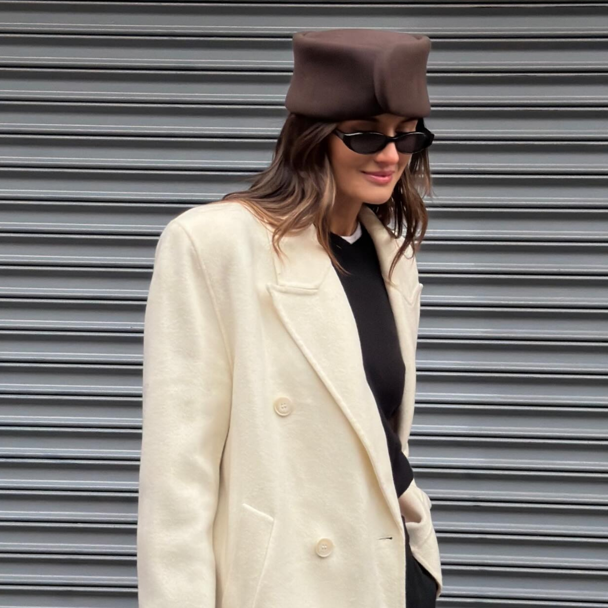 Coco Schiffer wears a brown pill-box hat, white coat, black sweater and black, oval sunglasses.
