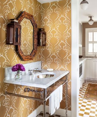 powder room with gold patterned wallpaper, marble sink, ornate wall mirror and vase of bright purple flowers