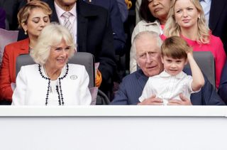 Queen Camilla in a white suit and King Charles sitting in chairs with Prince Louis sitting on The King's lap