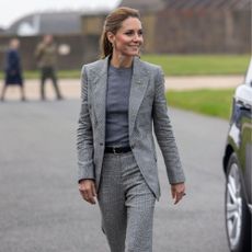 Kate Middleton walks outside wearing a tonal grey suit with navy suede heels and a black leather belt.