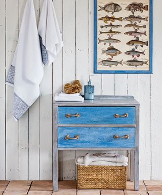 Shiplap walls with blue framed fish print, towels hanging, blue painted bathroom unit, baskets