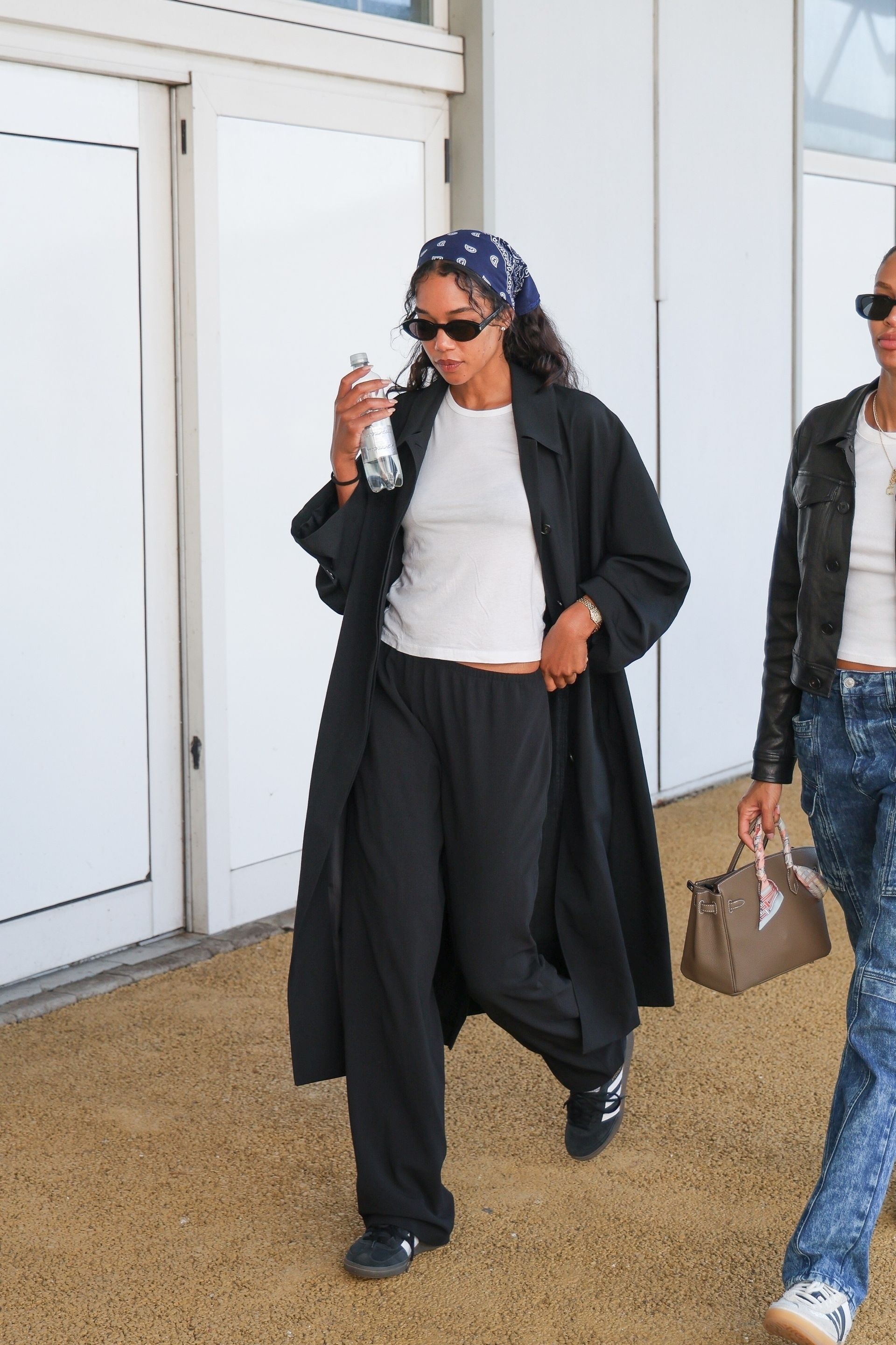 Laura Harrier walks through the airport wearing a white t-shirt, black pull-on trousers and black trousers. She layered with a black trench coat and wears a bandana over her hair.