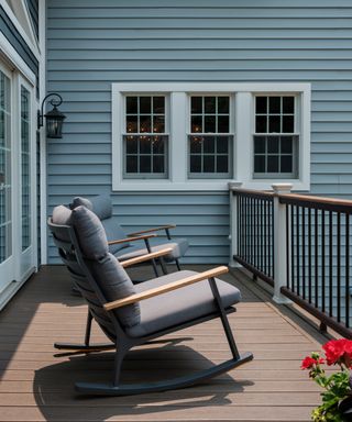 Gray rocking chair with plush cream cushions on a sunny deck with a blue exterior paneled wall in view behind