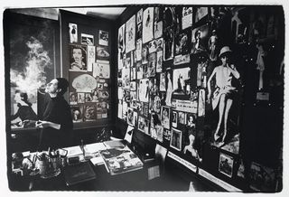 Black and white image with a man smoking in a room with a wall filled with pinned pictures on the wall behind