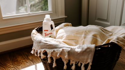 A wicker laundry basket with white linens and a bottle of detergent