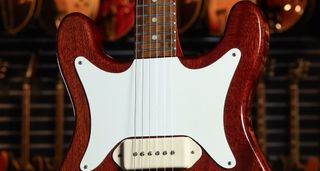 A close-up shot of a 1960 Epiphone Coronet in cherry red. This electric guitar has a lot of mojo with its white pickguard and cream pickup.