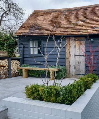 Painted she shed ideas, with a black clapboard garden room surrounded by gray paving, a wooden bench and neat hedges.