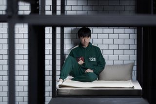 A man (Lee David) sits on a bunk bed against a white brick wall, while wearing a green tracksuit printed with 125, in 'Squid Game' season 2.