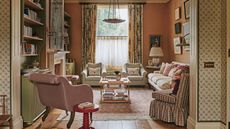 A warm, traditional sitting room with terracotta walls, floor-to-ceiling floral curtains, built-in olive green bookshelves, and a variety of patterned upholstered seating.