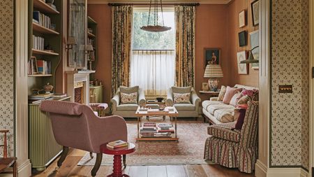A warm, traditional sitting room with terracotta walls, floor-to-ceiling floral curtains, built-in olive green bookshelves, and a variety of patterned upholstered seating.