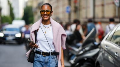  A guest wears orange sunglasses, gold earrings, a white halter-neck tank-top, gold chain pendant necklaces, a pale pink wool pullover, a black shiny leather shoulder bag from Givenchy, a black crossbody camera, a black shiny leather belt, blue faded denim large mom jeans pants, during the Bluemarble show, during Paris Fashion Week Menswear Spring Summer 2023, on June 21, 2022 in Paris, France. (Photo by Edward Berthelot/Getty Images )