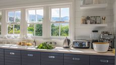 kitchen countertop with a chrome coffee maker, blender, and kettle on it. The counters are matte dark blue-gray and there are open crittal windows facing a beautiful woodland moutain landscape