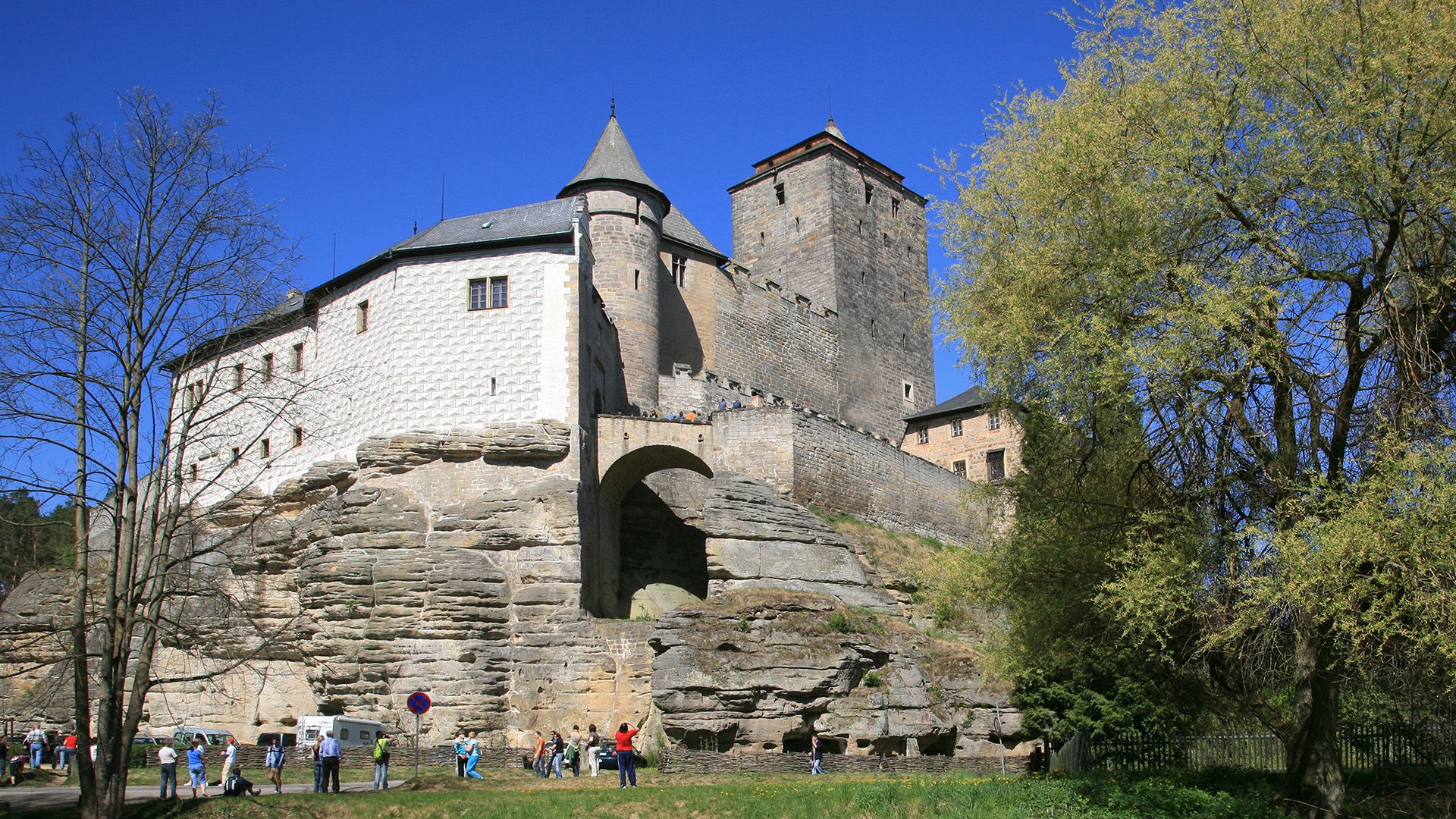 A view of a large stone wall with white brick buildings above as tourists walk below.
