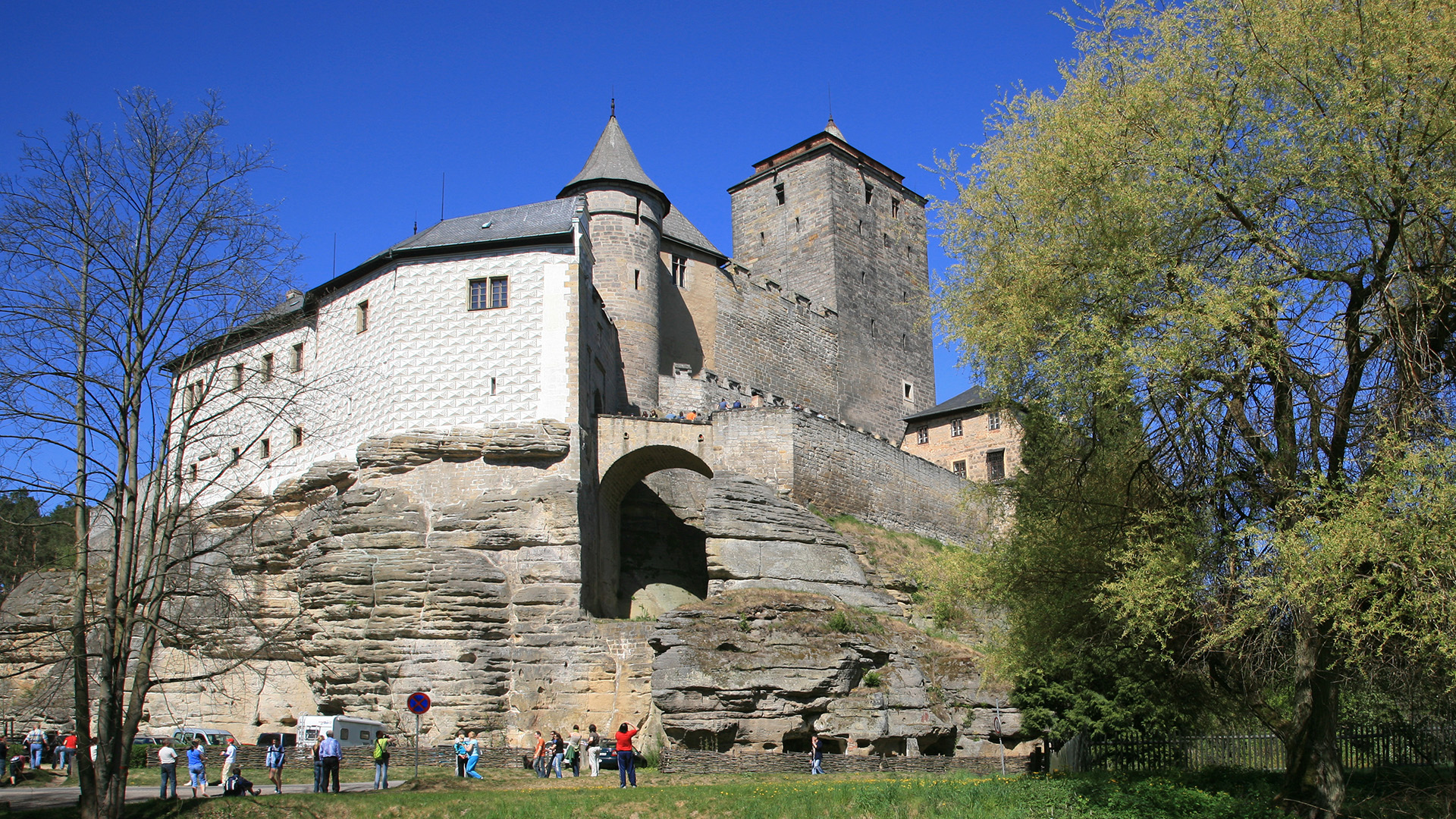 A view of a large stone wall with white brick buildings above as tourists walk below.