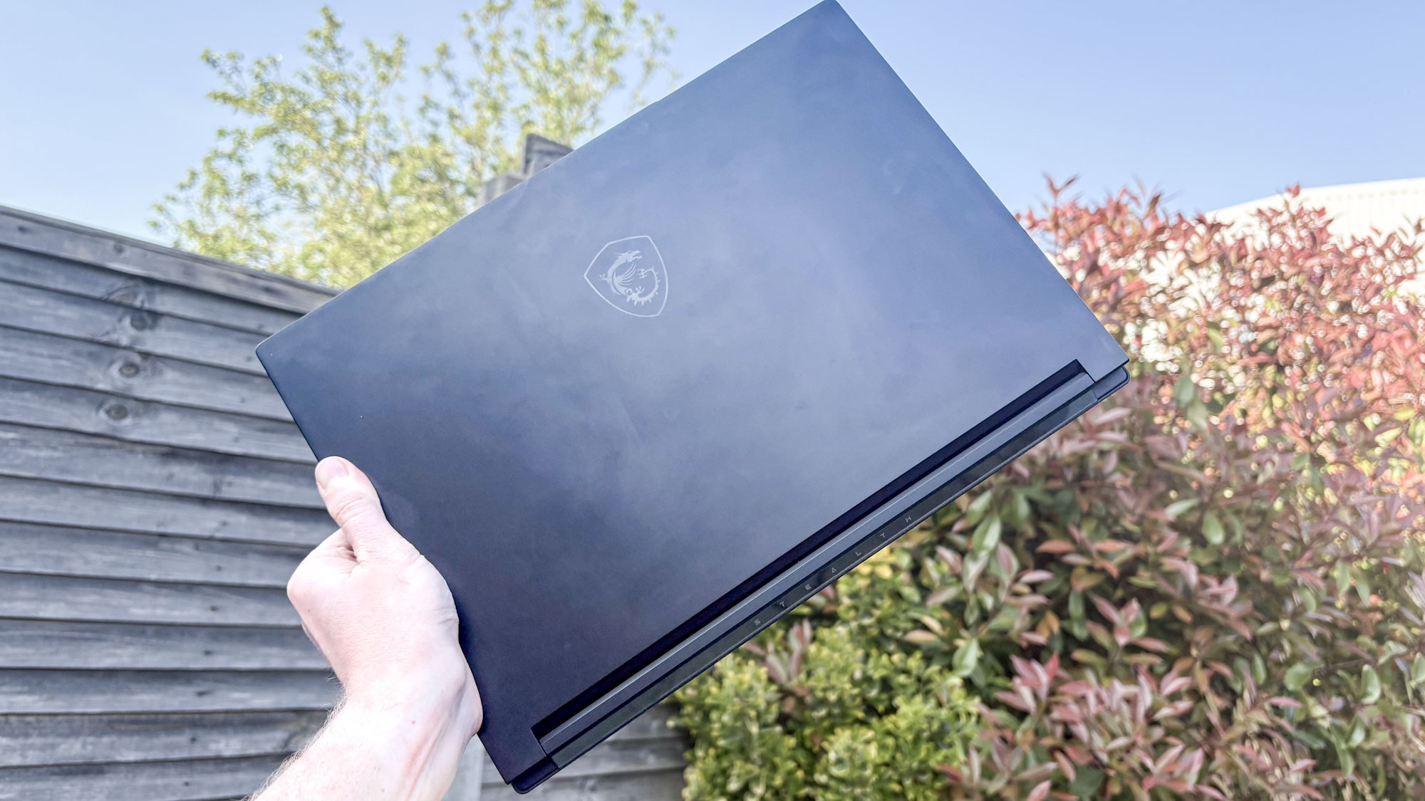MSI Stealth A16 AI+ closed in hand held up to sky with fence and bush in background