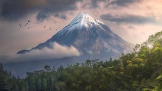 Snow-capped mountain rises above lush greenery, reflecting in a tranquil lake, with birds soaring in a moody, cloudy sky