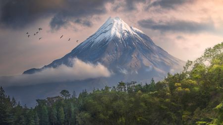 Snow-capped mountain rises above lush greenery, reflecting in a tranquil lake, with birds soaring in a moody, cloudy sky