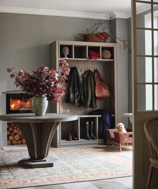 winter decor ideas, grey boot room with open shelving/hanging cabinet, large wooden round table, patterned rug, log burner, basket, large vase of dyed eucalyptus
