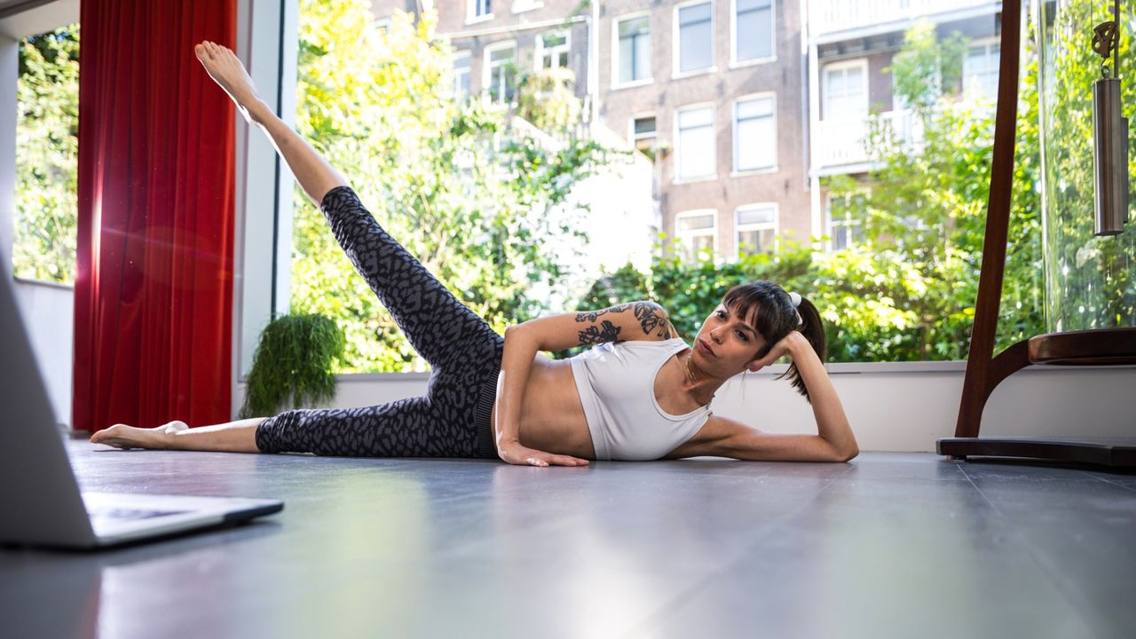 woman lying on her side performing a leg lift in front of a laptop, facing the camera. Behind her is a large window looking out to trees and flats. 