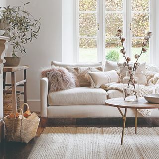 A living room with a cream sofa and multiple textured cushions and a knitted throw on top and a round wooden coffee table in front of it