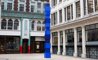 A tall vertical structure, created using square blocks in different shades of blue, in an open area in front of a building