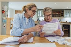 A senior woman and her mature daughter at home in Seghill, Northumberland. The senior woman is staying with her daughter for support and care as she has dementia. They are sat at the dining room table where they're looking through financial bills, looking concerned.