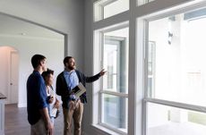 Confident male real estate agent gestures toward a beautiful view out the window of a new home. Interested potential homeowners attentively listen to the real estate agent.