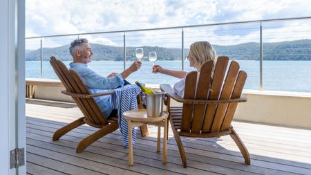 A retired couple toast each other with wine while sitting on a deck with a water view.