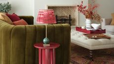 a living room with a green velvet sofa and large fireplace with artwork on, a table lamp is on a table in the foreground