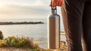 Woman holding stainless steel water bottle looking out at view across the sea on a hike, avoiding some of the most common hiking mistakes