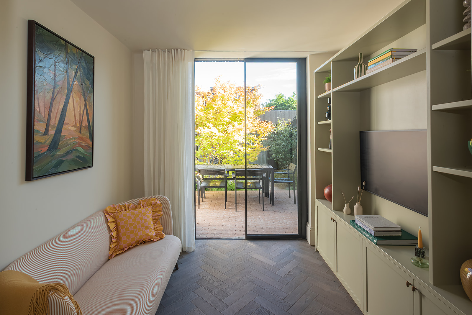a modern living room with built-in shelving and a TV, sofa opposite with artwork, sofa with two yellow ruffle cushions, parquetry floors, and sliding glass doors leading to outside