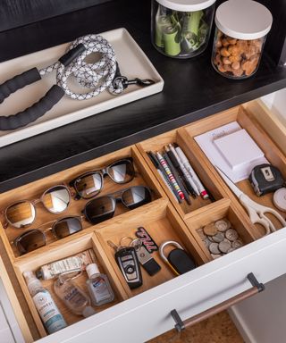 An open wooden drawer with sunglasses, keys, notepads, and hand sanitizer inside it