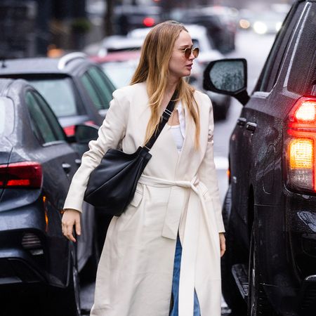 Jennifer Lawrence is in the street while walking and wearing a trench coat, jeans, black messenger bag, and gray Adidas Handball Spezial shoes.