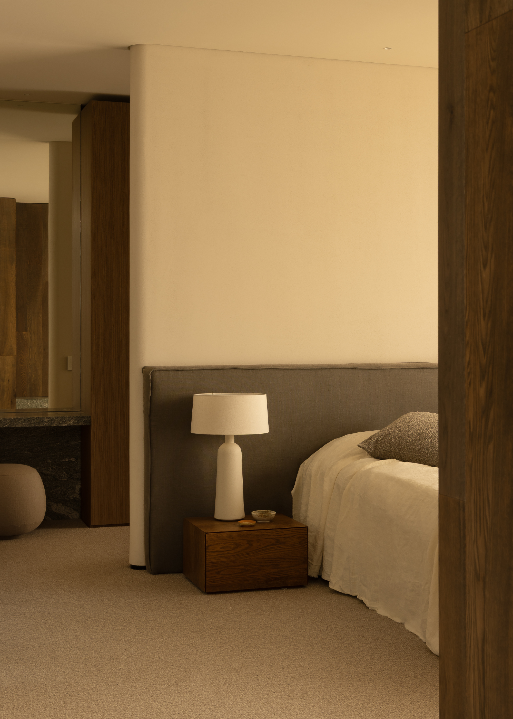 An off-white bedroom with a gray extended headboard, a white bedscape, a low wood nightstand and a white table lamp