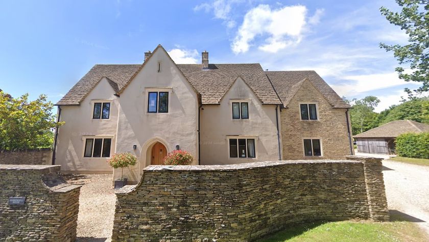 A white house with a stone front wall and a log barn to the side with a long driveway